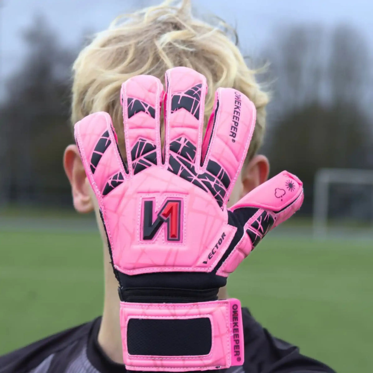 Pink youth goalkeeper gloves with black grip pattern, worn on a player's hand on a soccer field