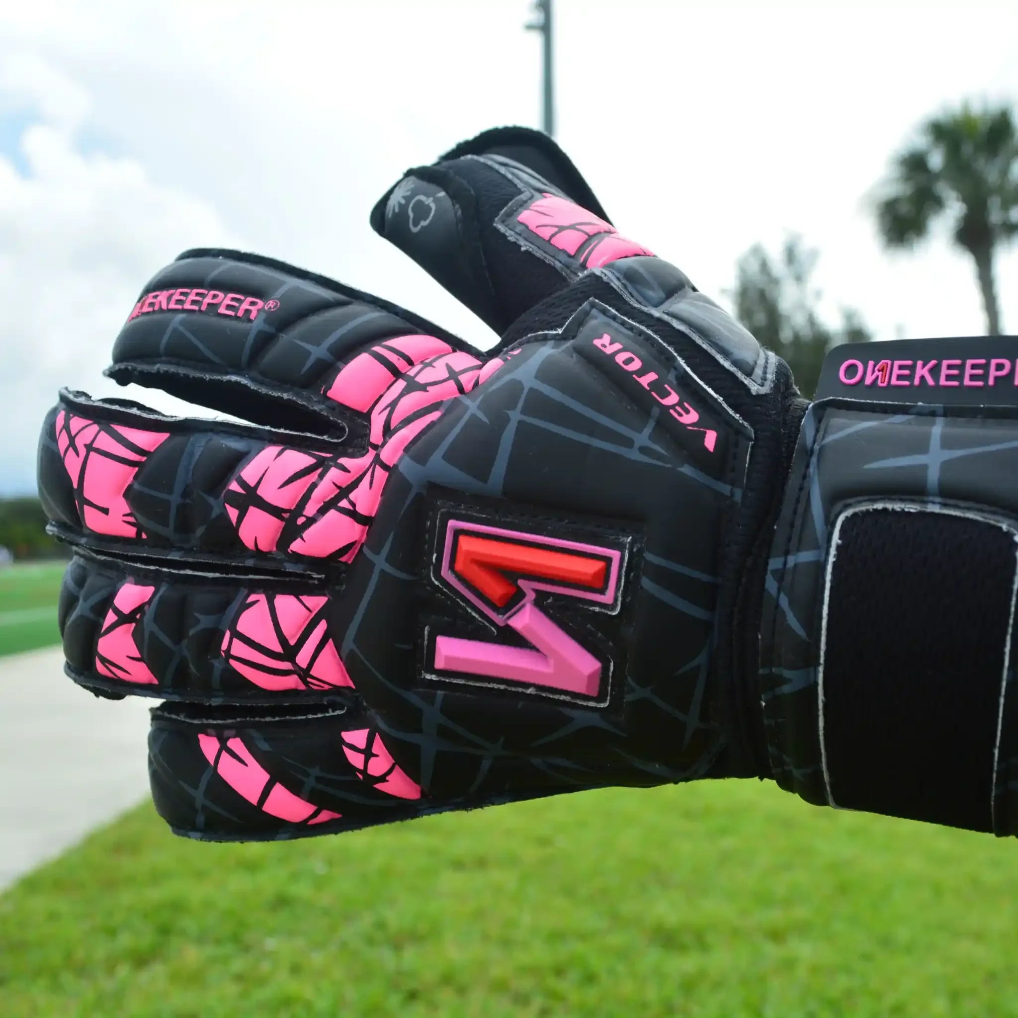 ONEKEEPER Vector Evolution Black and pink soccer goalkeeper gloves with visible branding on a grassy background.
