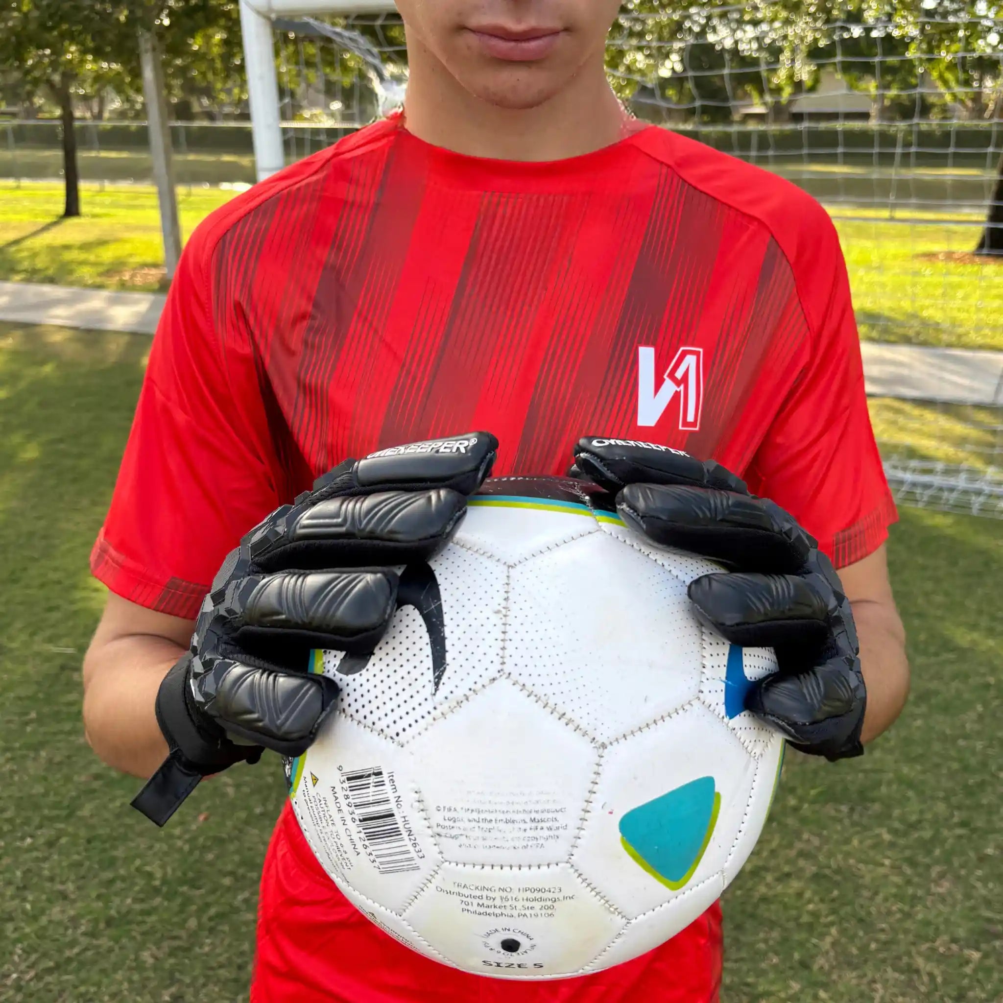 Person wearing a red sports jersey and a ONEKEEPER ACE black glove holding a soccer ball on a grassy field.