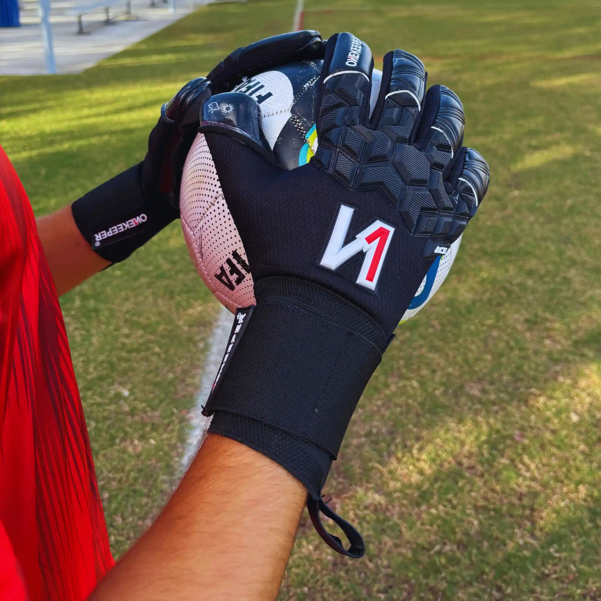 Person wearing a onekeeper ace black sports glove with a red 'N' on a grassy field