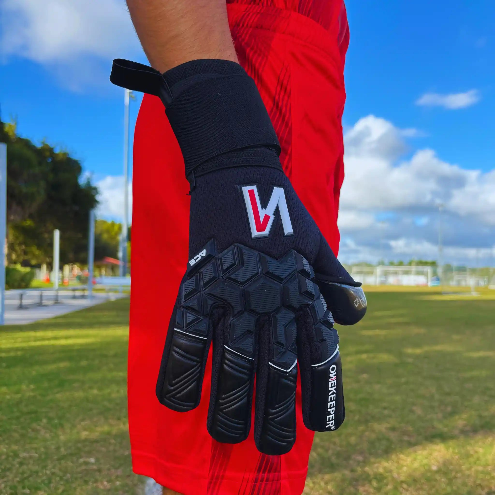 ONEKEEPER ACE Black glove with a logo on a person wearing red pants, standing on a grassy field.