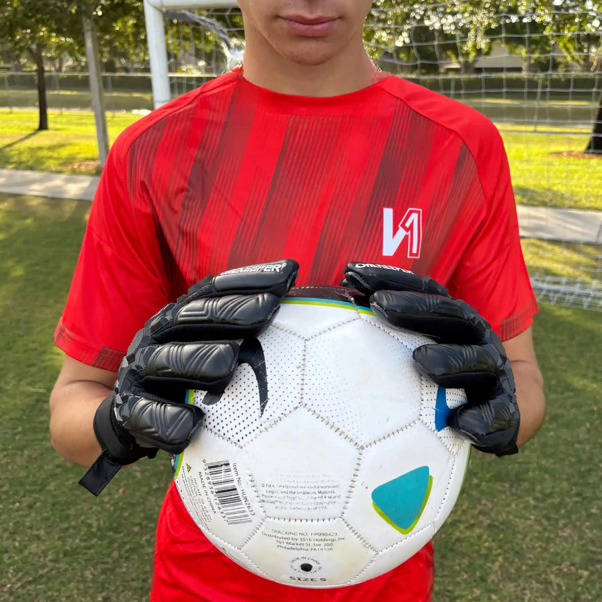 Person wearing a red sports jersey and onekeeper ace black gloves holding a soccer ball on a grassy field.