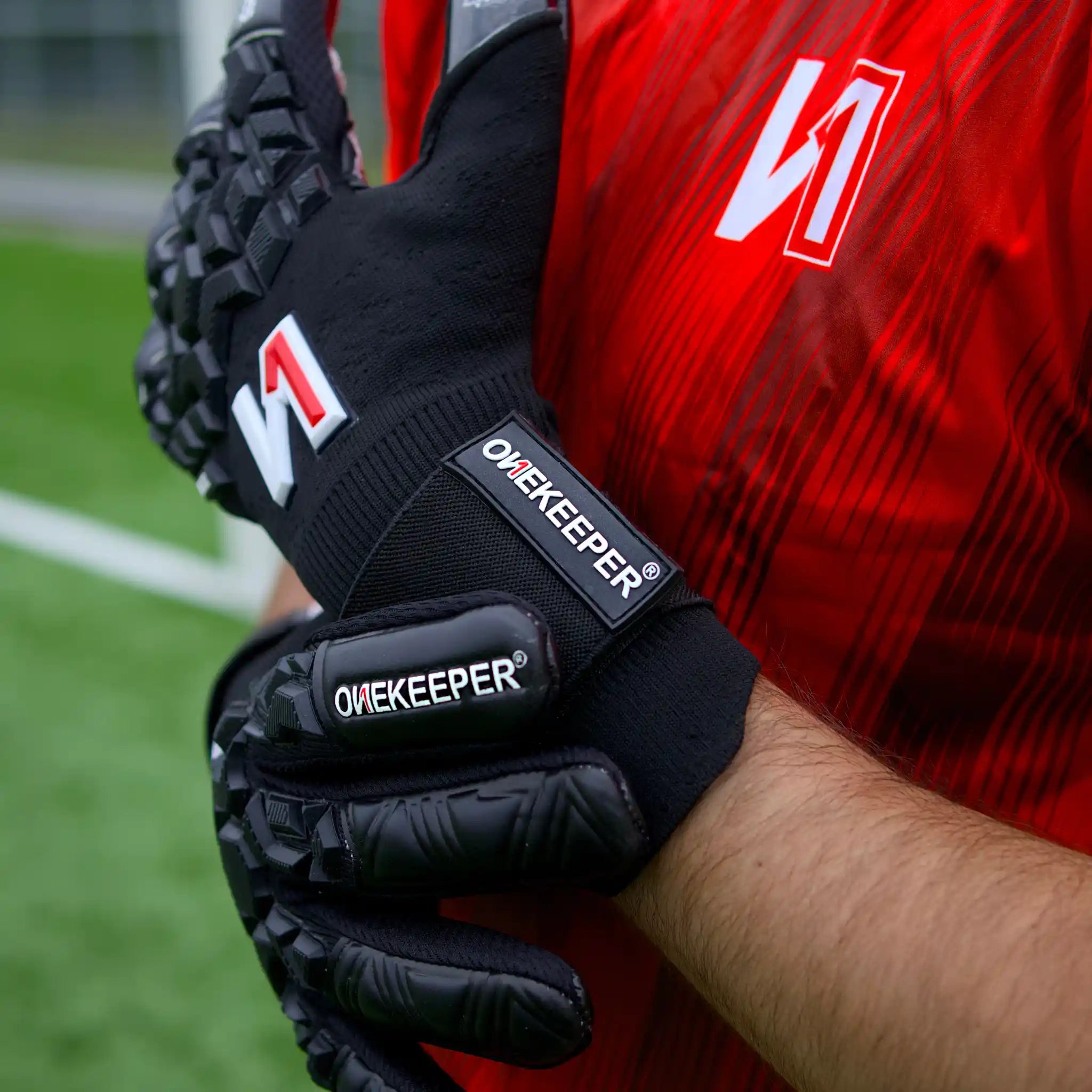 Person wearing A onekeeper ACE black goalkeeper gloves with 'Onekeeper' branding on a sports field.