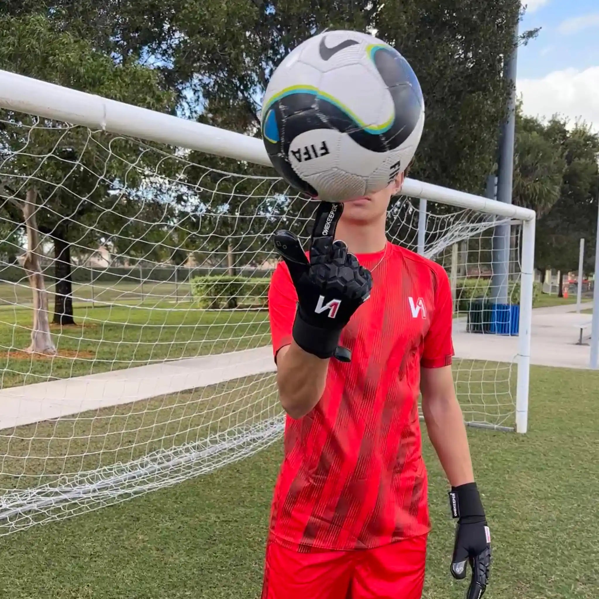 Person in red soccer uniform holding a soccer ball in front of a goalpost wearing an ONEKEEPER ACE Black glove.