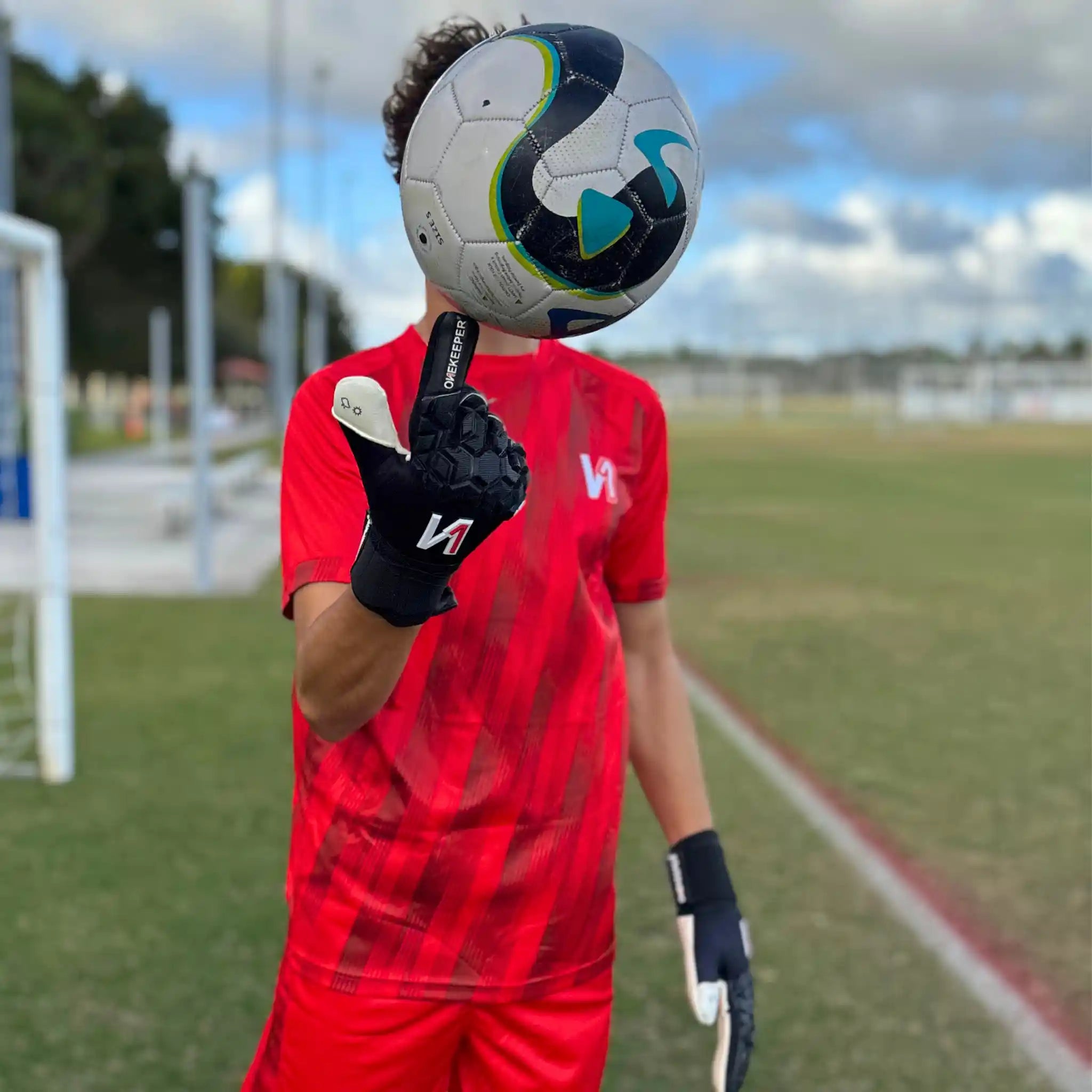 Person in red sports uniform with a soccer ball on a sports field wearing a ONEKEEPER ACE Black and White goalkeeper gloves