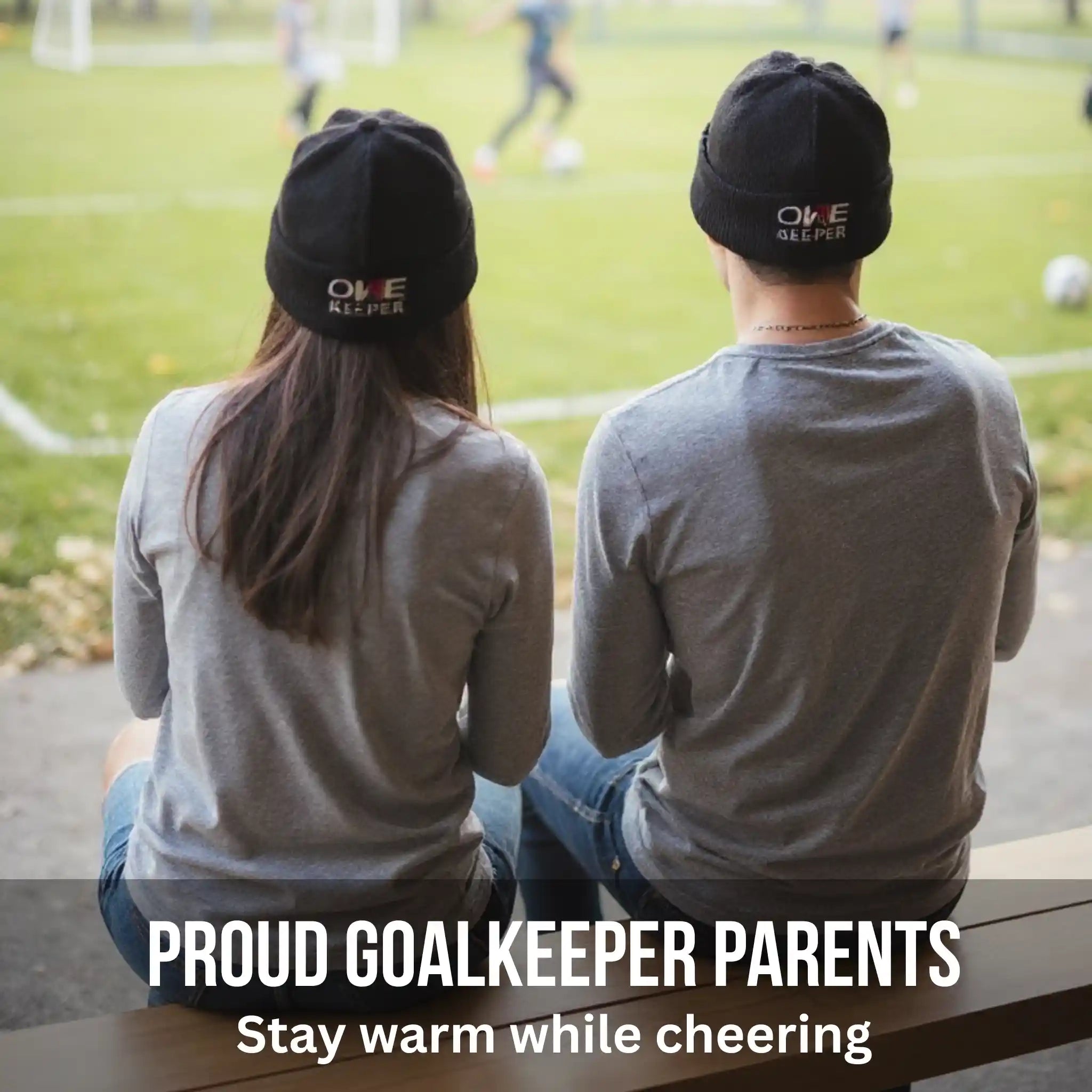 Two people wearing black beanies with 'ONE KEEPER' text, sitting on a bench with a soccer field in the background.