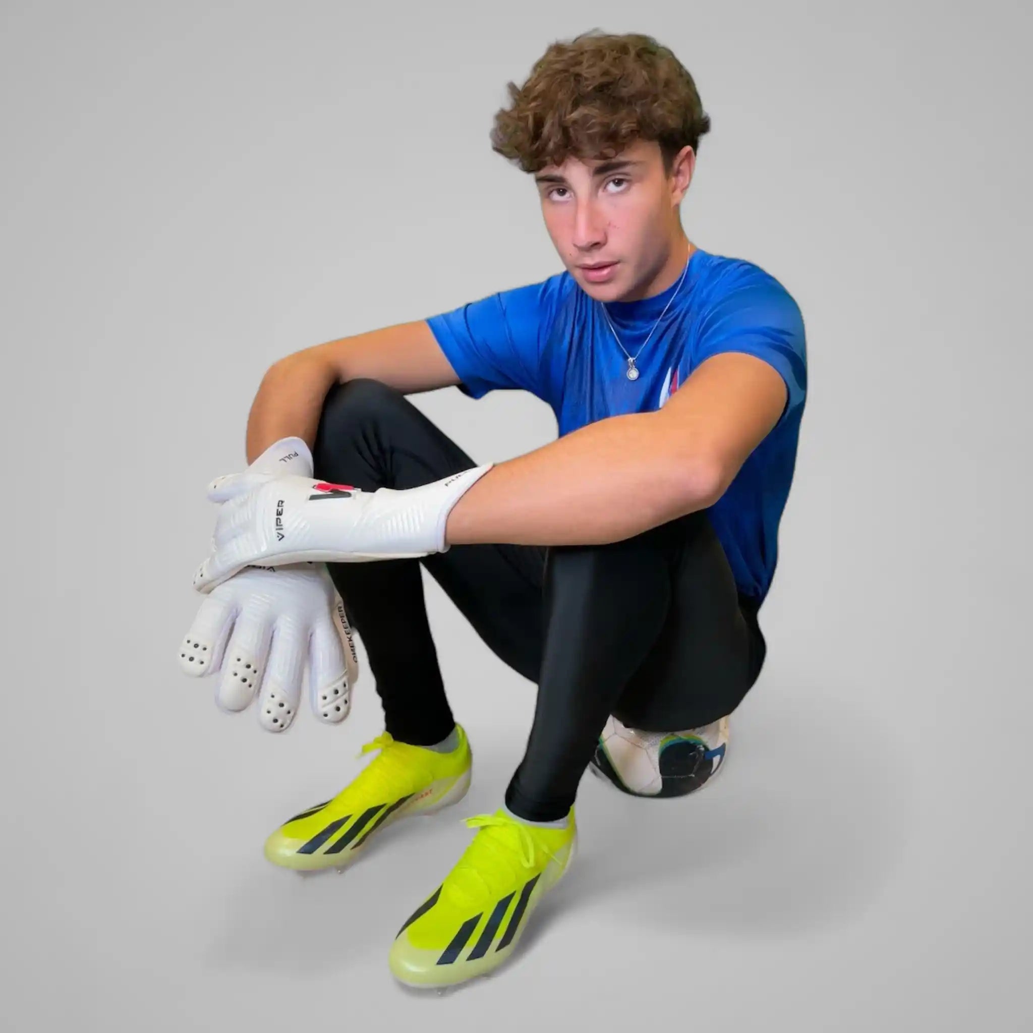 Person wearing a blue sports outfit with black goalkeeper compressions leggings, white gloves, and green soccer cleats on a gray background