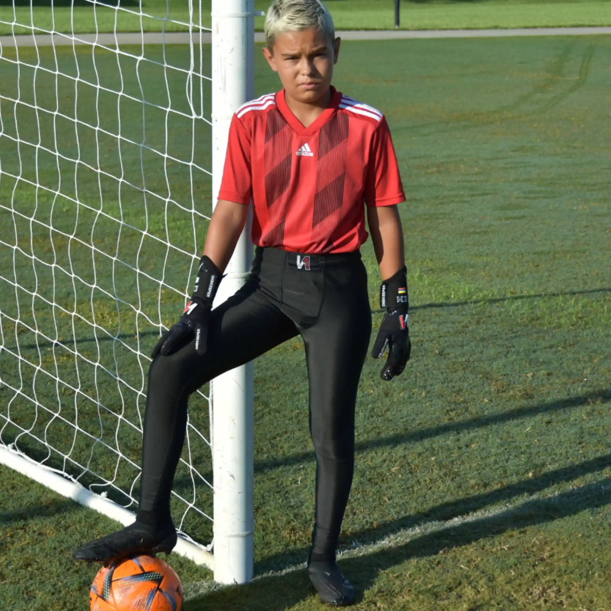 A youth goalkeeper in a red soccer jersey and black compression leggings is standing on a grass field with a soccer ball.