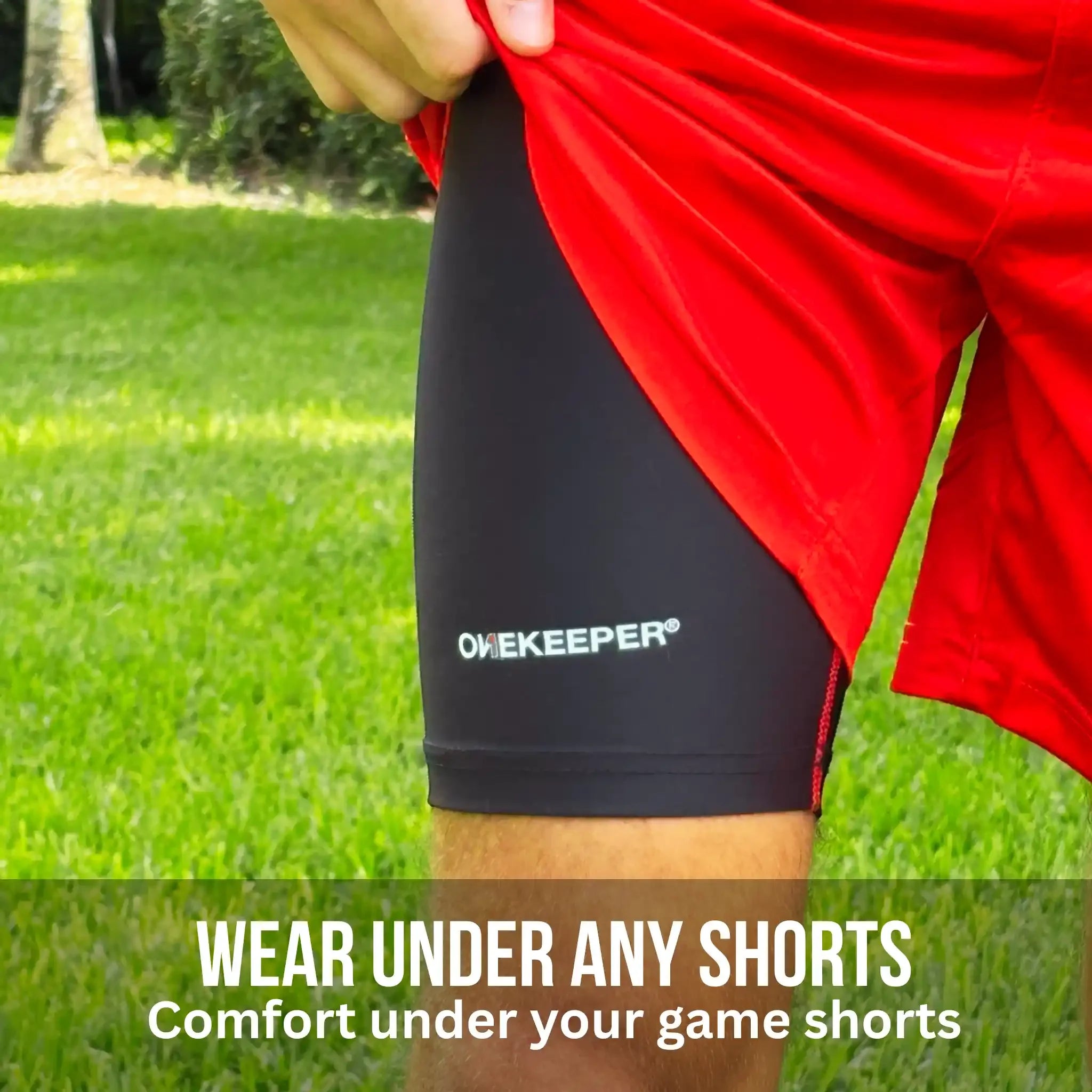 Person wearing onekeeper compression black shorts with ONEKEEPEER logo, holding a red garment on grassy background