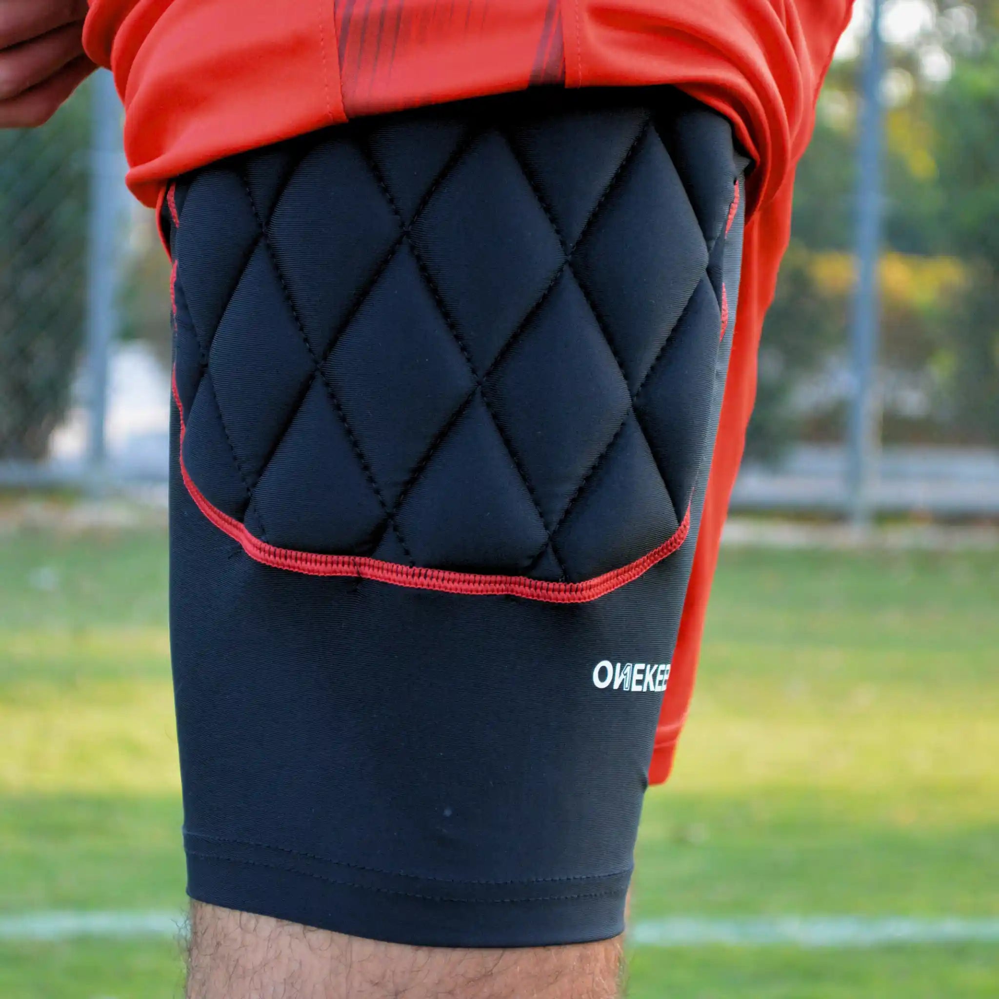 Black quilted knee pad with red trim on a person wearing a red sports outfit, on a grassy field.