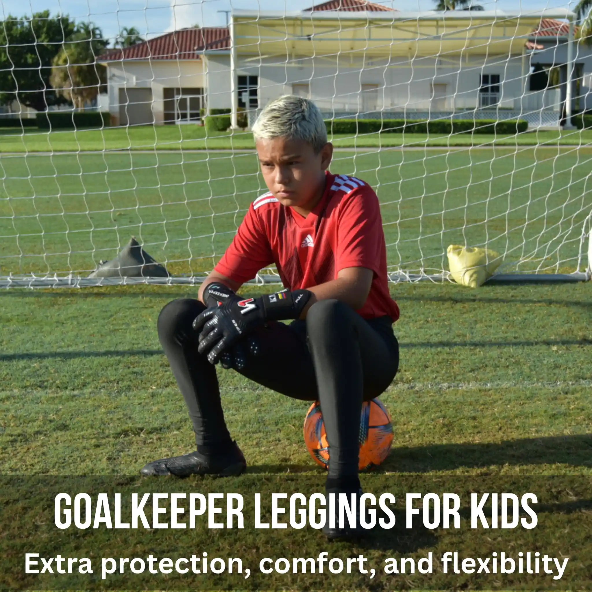 Child in red soccer jersey and black goalkeeper leggings sitting on a soccer ball in front of a goal.