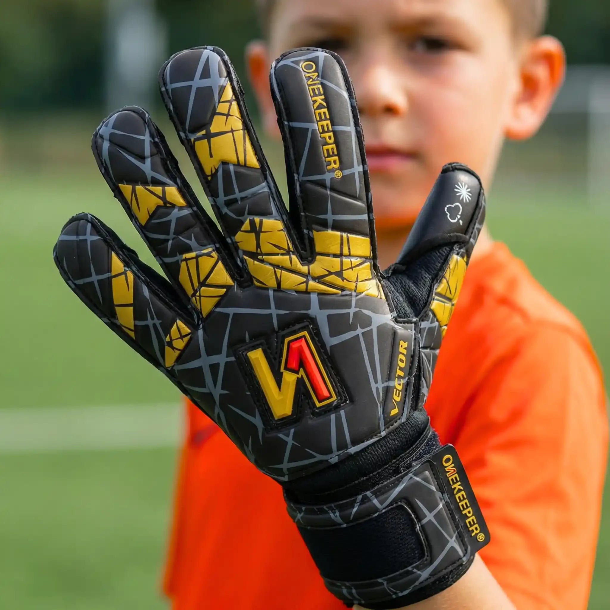 onekeeper vector evolution black and gold Goalkeeper glove with geometric pattern and 'ONEKEEPER' branding held by a child on a blurred sports field background.