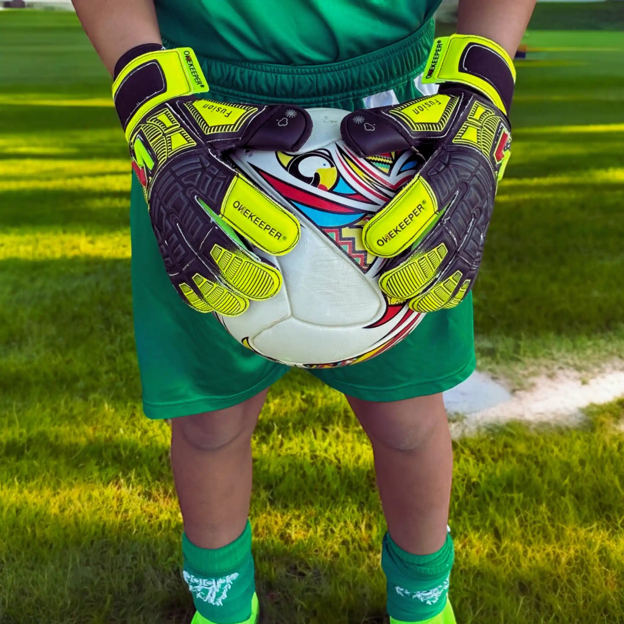 Person wearing a onekeeper fusion green goalkeeper gloves and shorts holding a colorful soccer ball on a grass field.