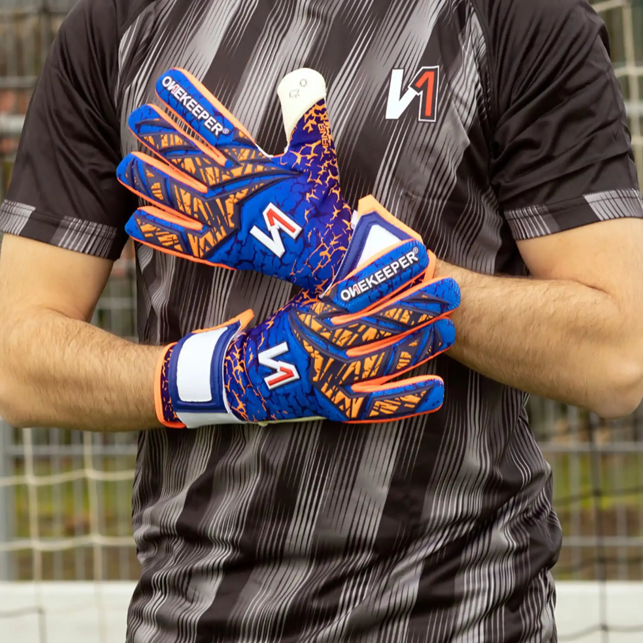 Soccer goalkeeper wearing a onekepeer mana blue and orange goalie gloves with geometric patterns, standing on field.