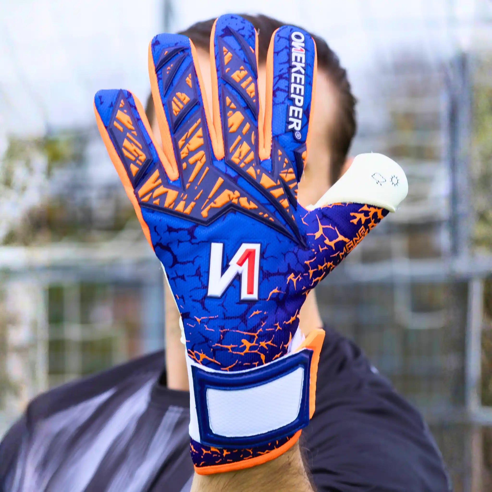 A close-up of a onekeeper manage blue and orange goalkeeper glove worn on a hand, with the back of the glove facing the camera. The glove features a blue and orange patterned design with white accents and the brand name 'ONEKEEPER' printed vertically on the finger.
