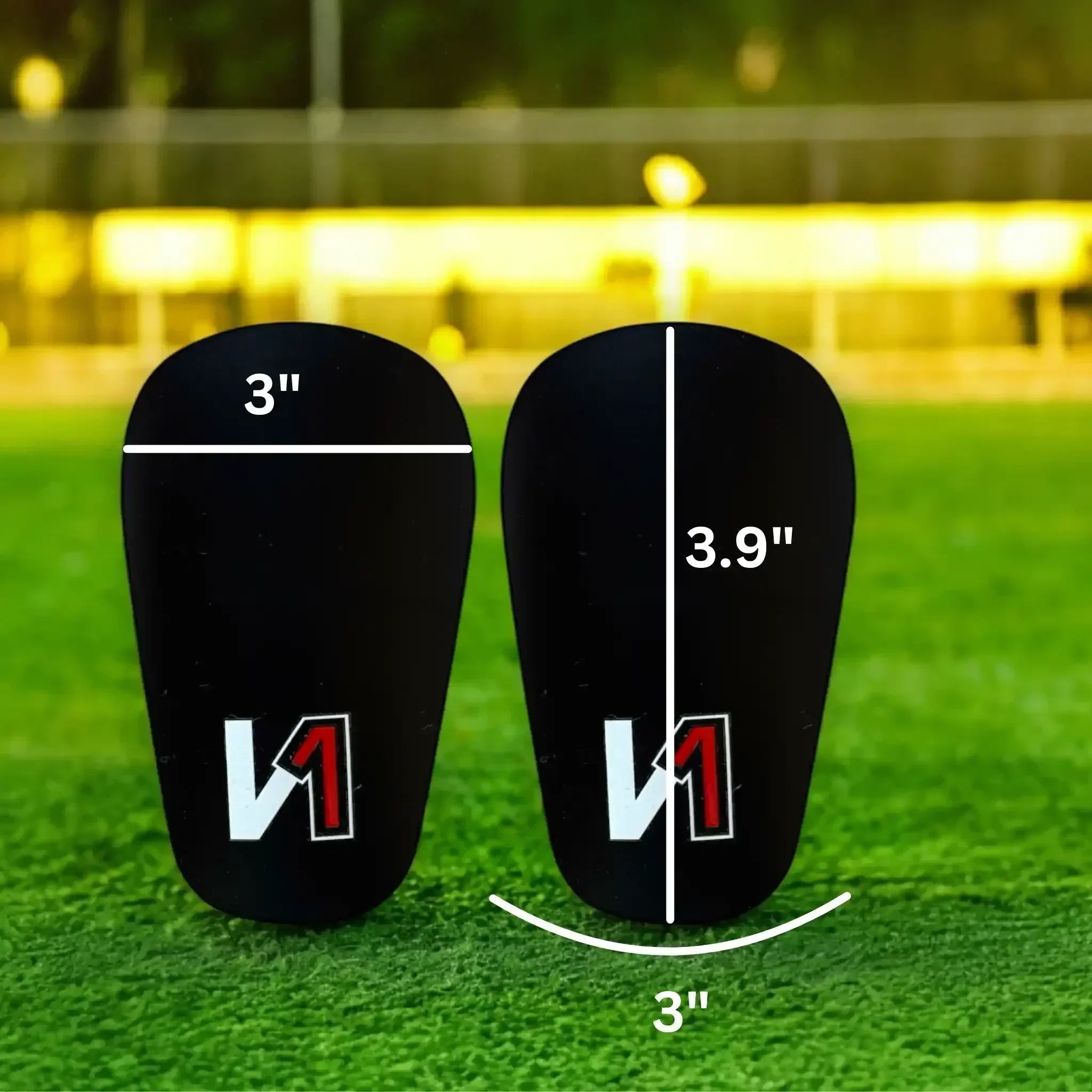 Black mini shin guards with measurement lines, 3 inches by 3.9 inches, on a soccer field