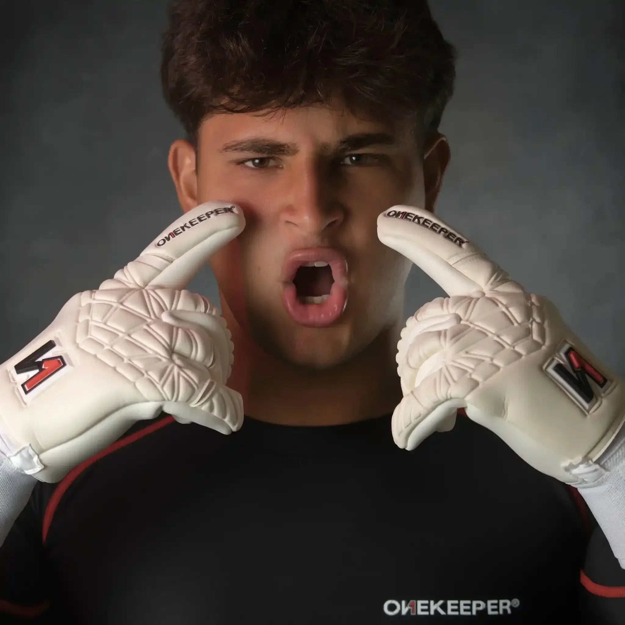 Soccer goalkeeper wearing onekeeper philipp kohn white textured gloves and black shirt, pointing at his face