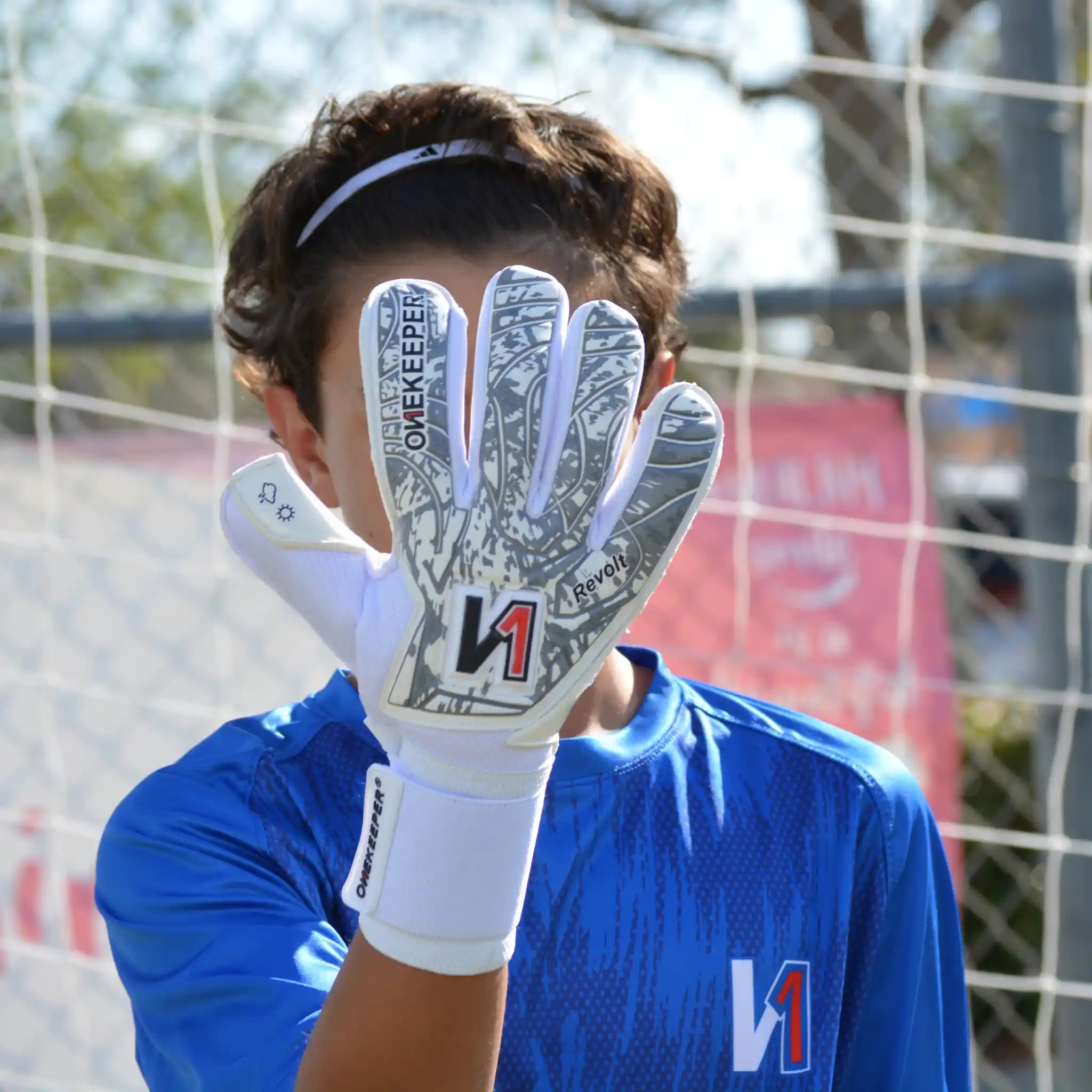 Person wearing a onekeeper revolt white goalkeeper glove with a visible brand logo, standing in front of a goalpost.