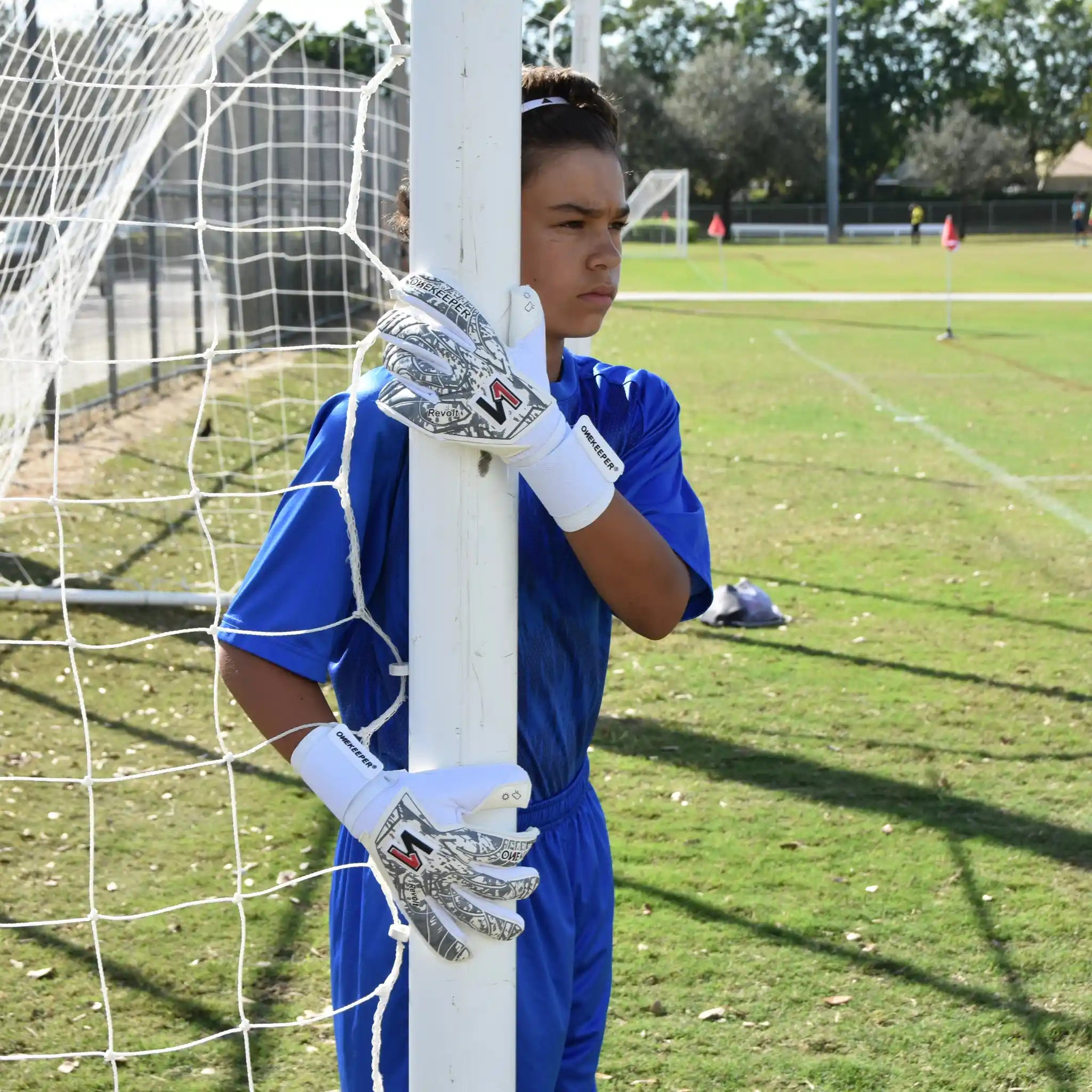 Person in blue soccer uniform with a onekeeper revolt white gloves standing behind a goalpost on a soccer field.