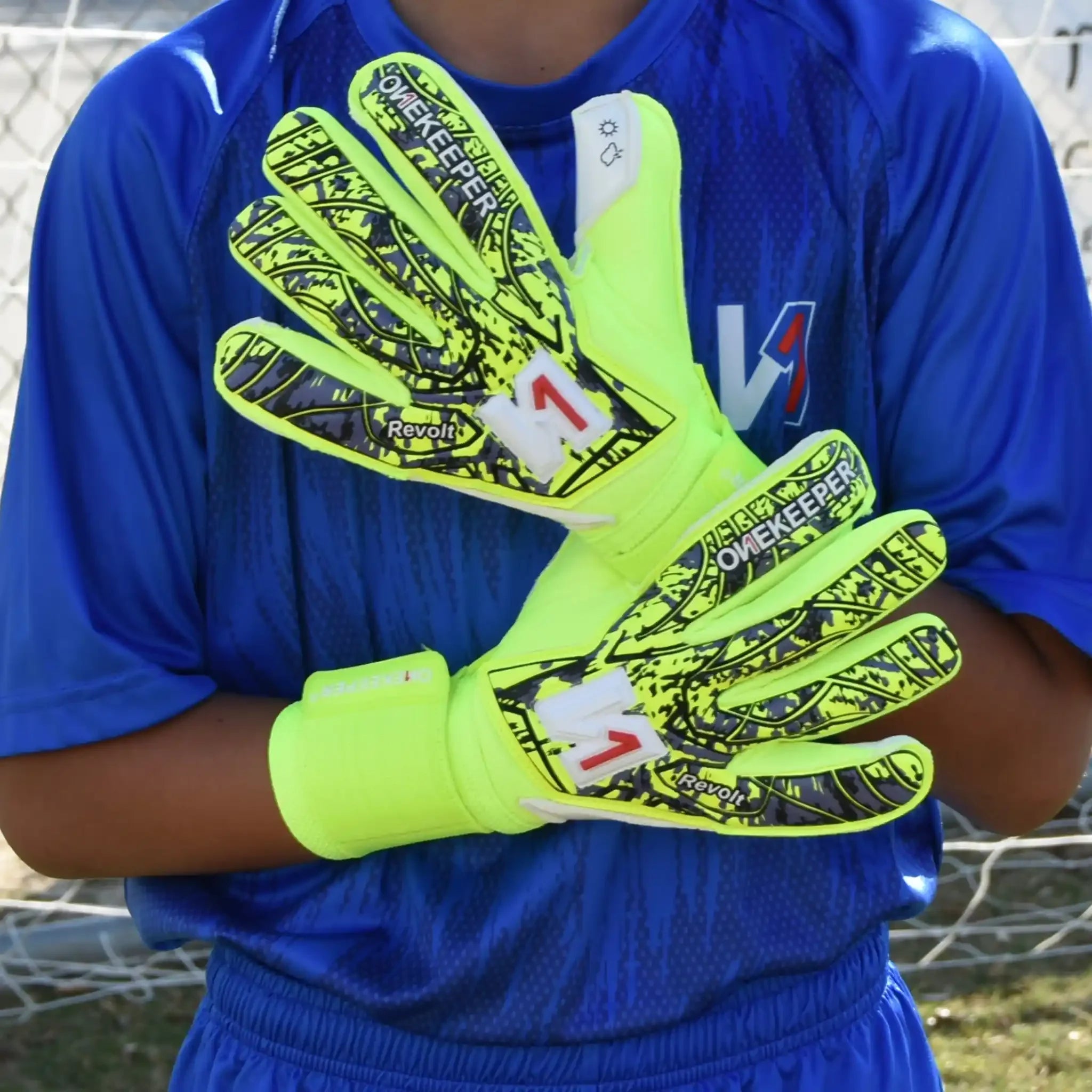 Person wearing a onekeeper revolt fluo yellow goalkeeper gloves with black patterns, holding a soccer ball, against a blue sports jersey.