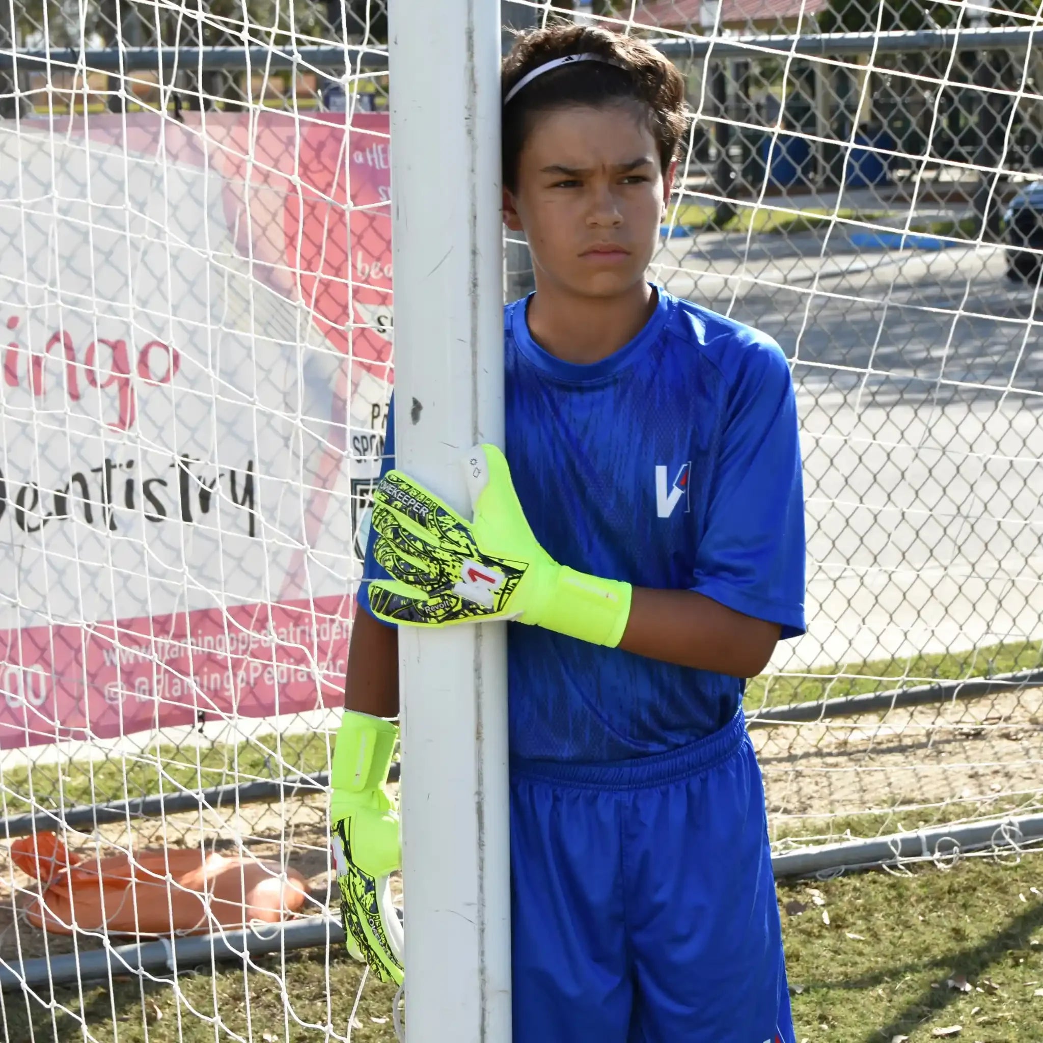 Person in blue soccer goalkeeper uniform with onekeeper revolt fluo yellow gloves leaning against a goalpost.