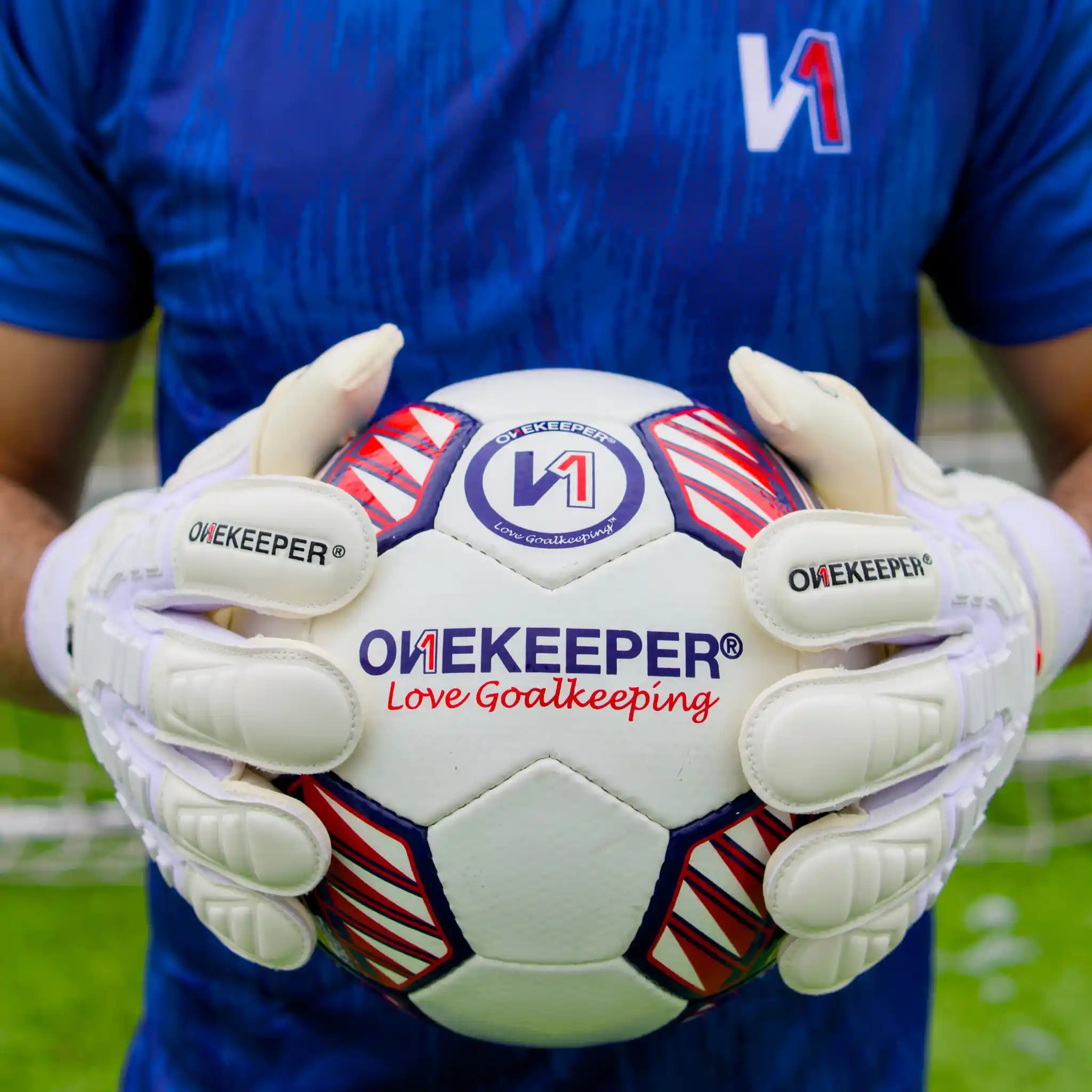 Person holding a soccer ball with 'ONEKEEPERS' branding, wearing gloves on a grassy field.