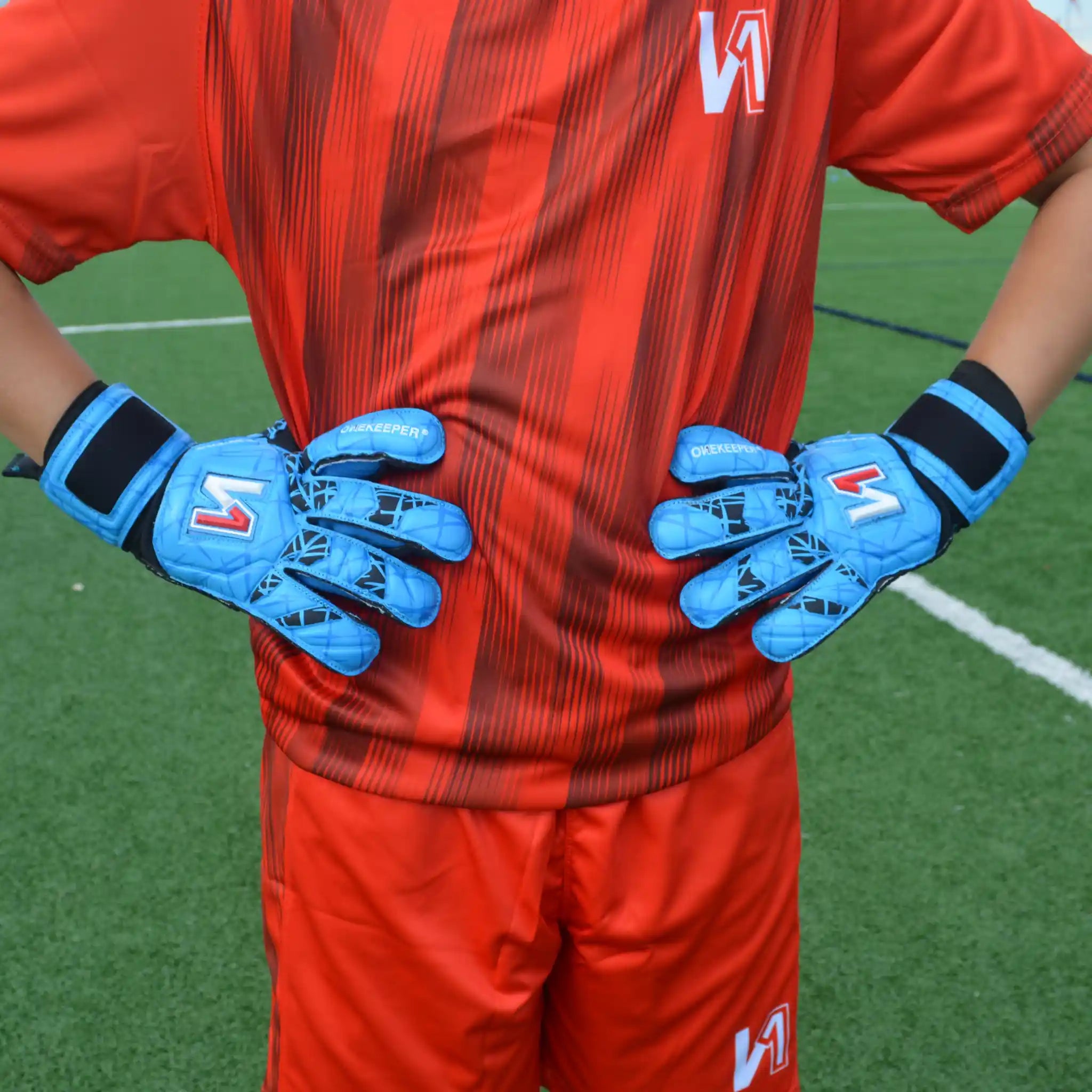 Person wearing red sports uniform and ONEKEEPER Vector Evolution blue gloves with a logo on a grass field