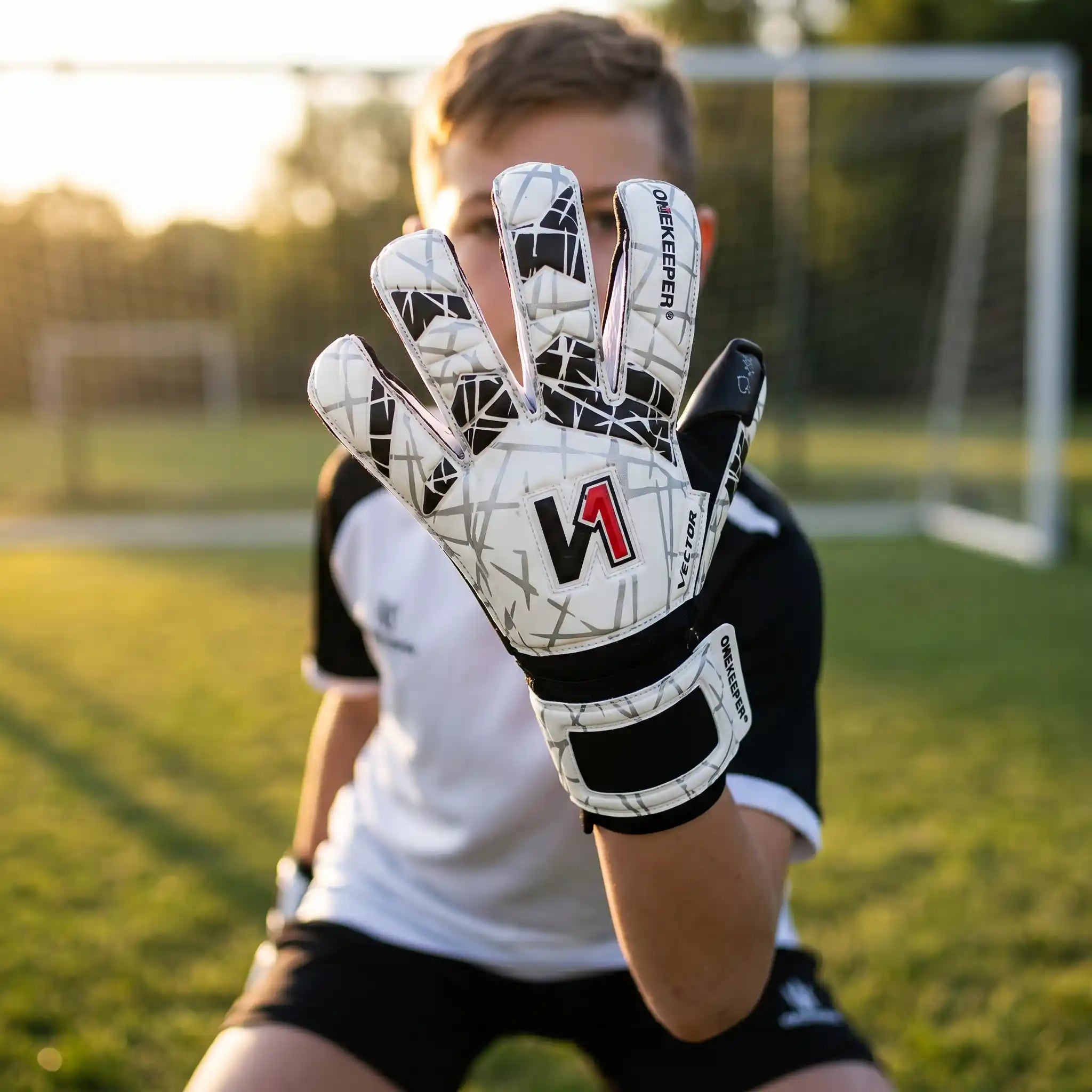 Person on a soccer field wearing a onekeeper vector evolution white and black goalkeeper glove with black patterns and a red 'V1' logo.