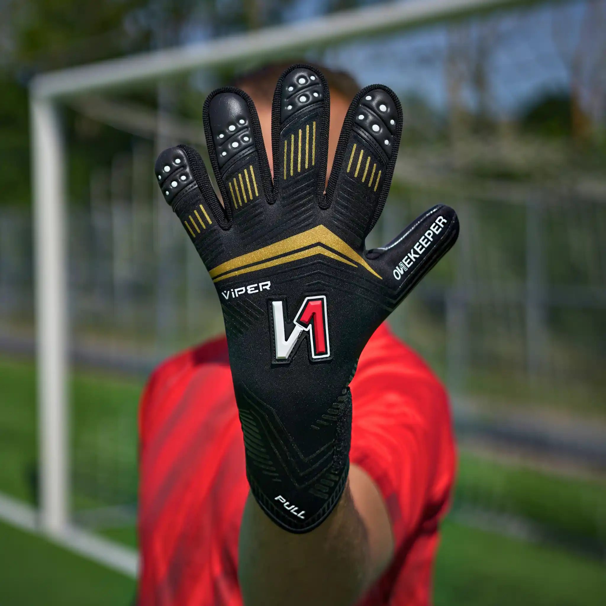 A onekeeper viper black and gold soccer goalkeeper glove with gold and white accents, featuring the brand name 'Viper' and model 'Keeper', held up in front of a soccer field background.
