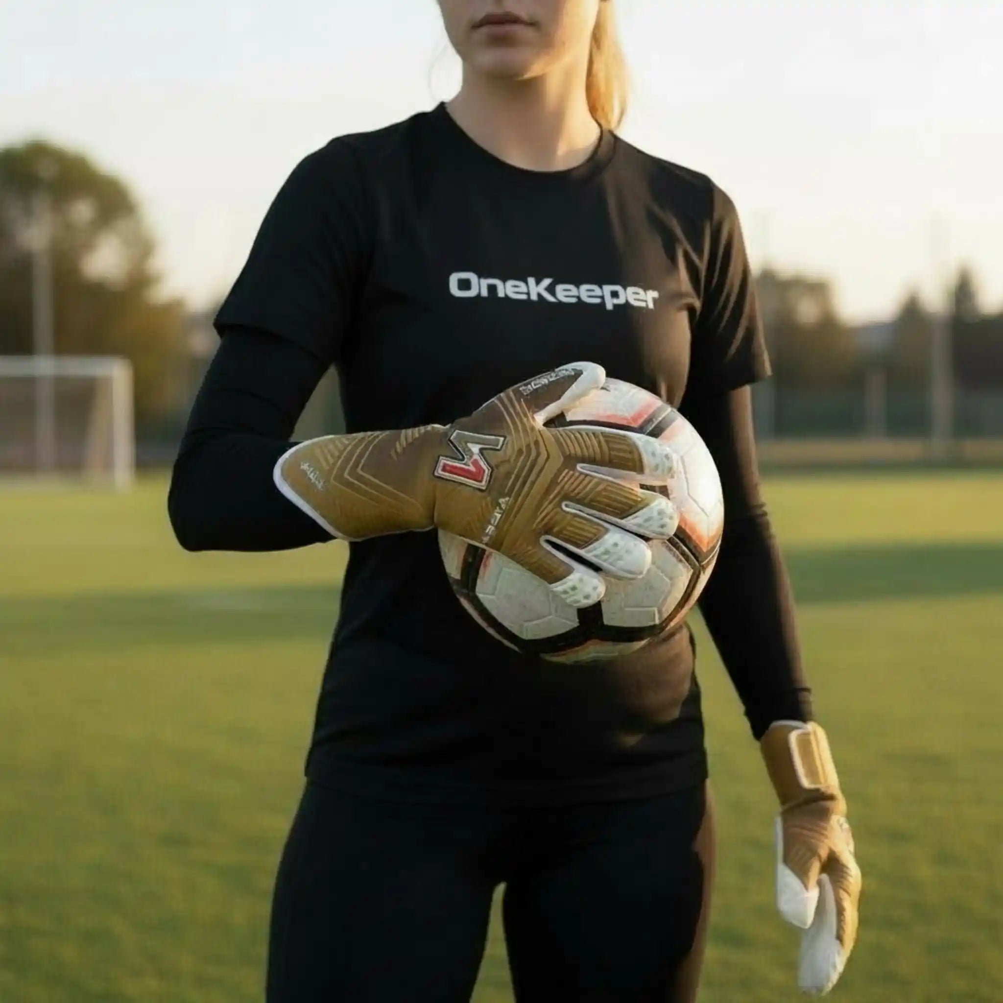 Person wearing a black 'OneKeeper' shirt holding a onekeeper viper gold glove on a grassy field.