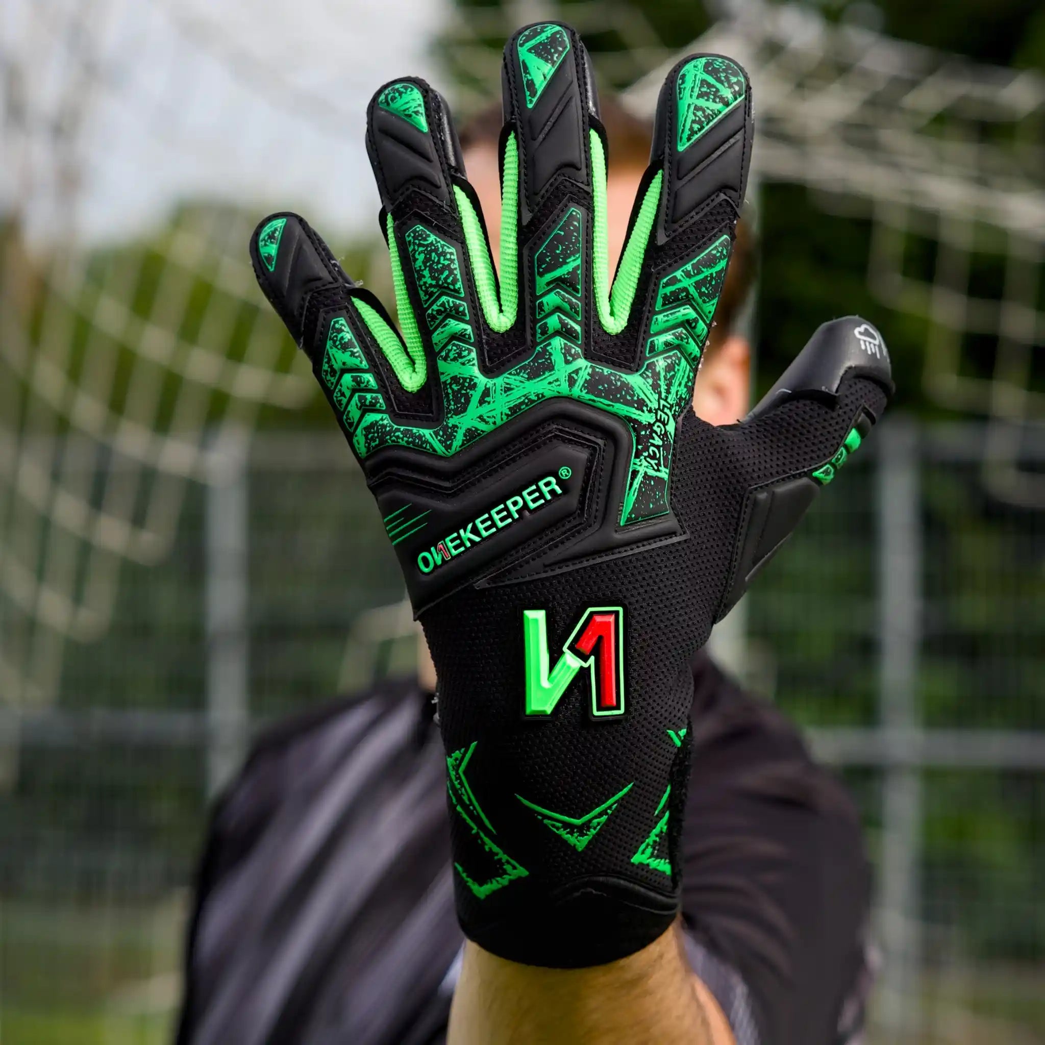A close-up of a onekeeper legacy black and green goalkeeper glove held up in front of a blurred soccer goal background. The glove is primarily black with bright neon green accents and patterns, featuring the brand name 'ONEKEEPER' and a stylized 'V1' logo.