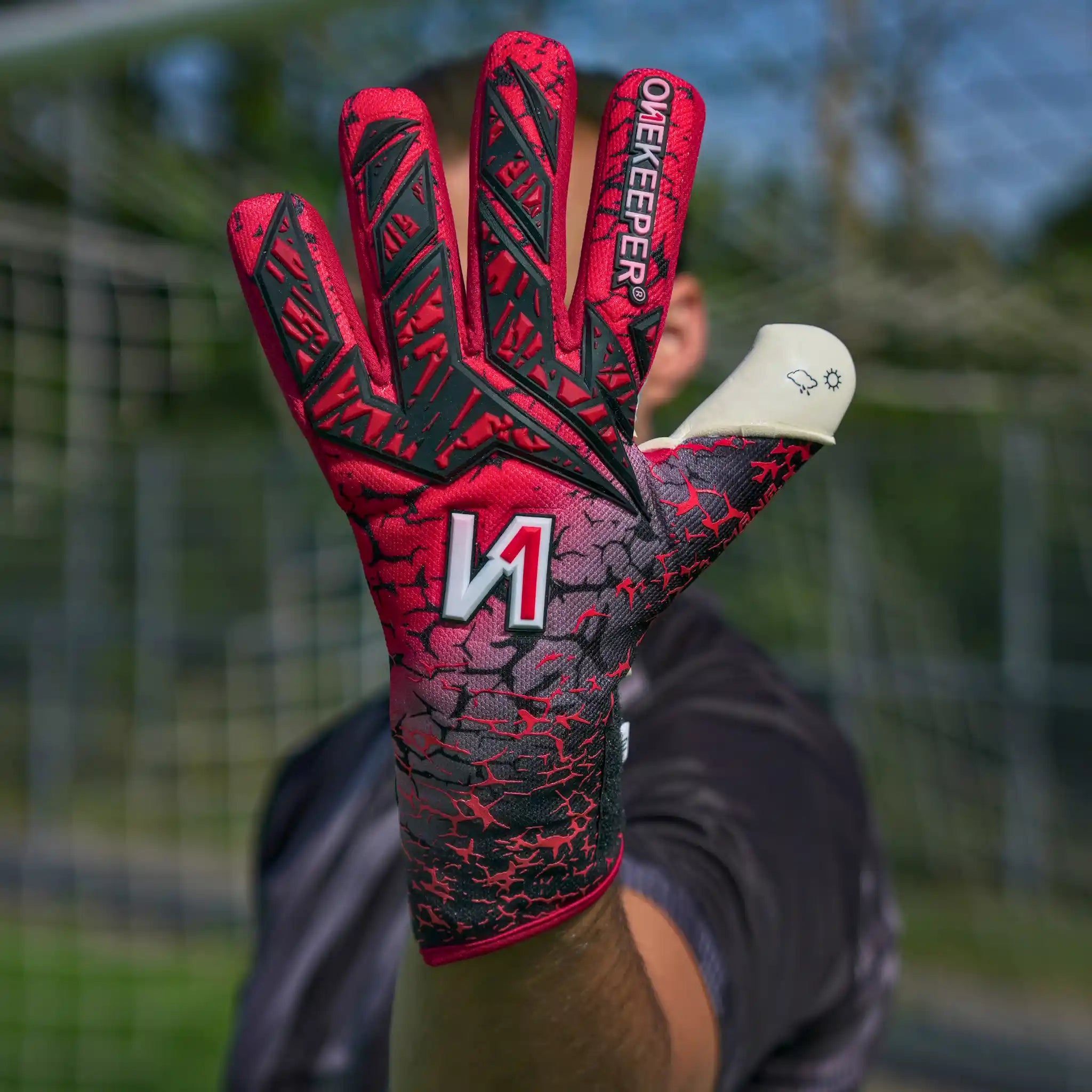 A close-up of a onekeeper mana red goalkeeper glove held up toward the camera, with a blurred person and outdoor field background. The glove is red and black with a cracked pattern, a large white and red 'M' logo on the back, and the word 'ONEKEEPER' printed vertically on the finger.