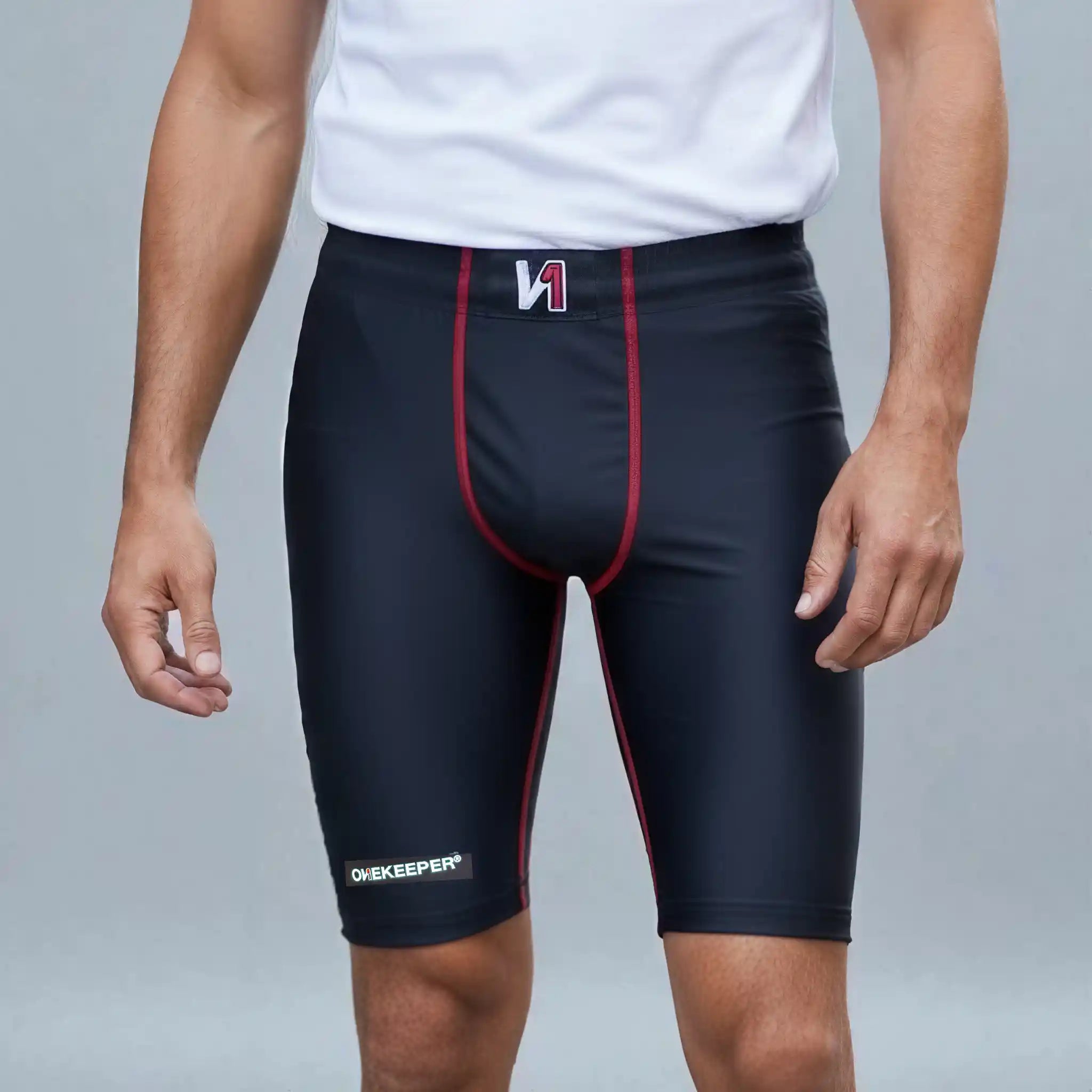 A man wearing a onekeeper compression shorts black athletic compression shorts with red piping and a white logo on the waistband, paired with a white t-shirt against a gray studio background.