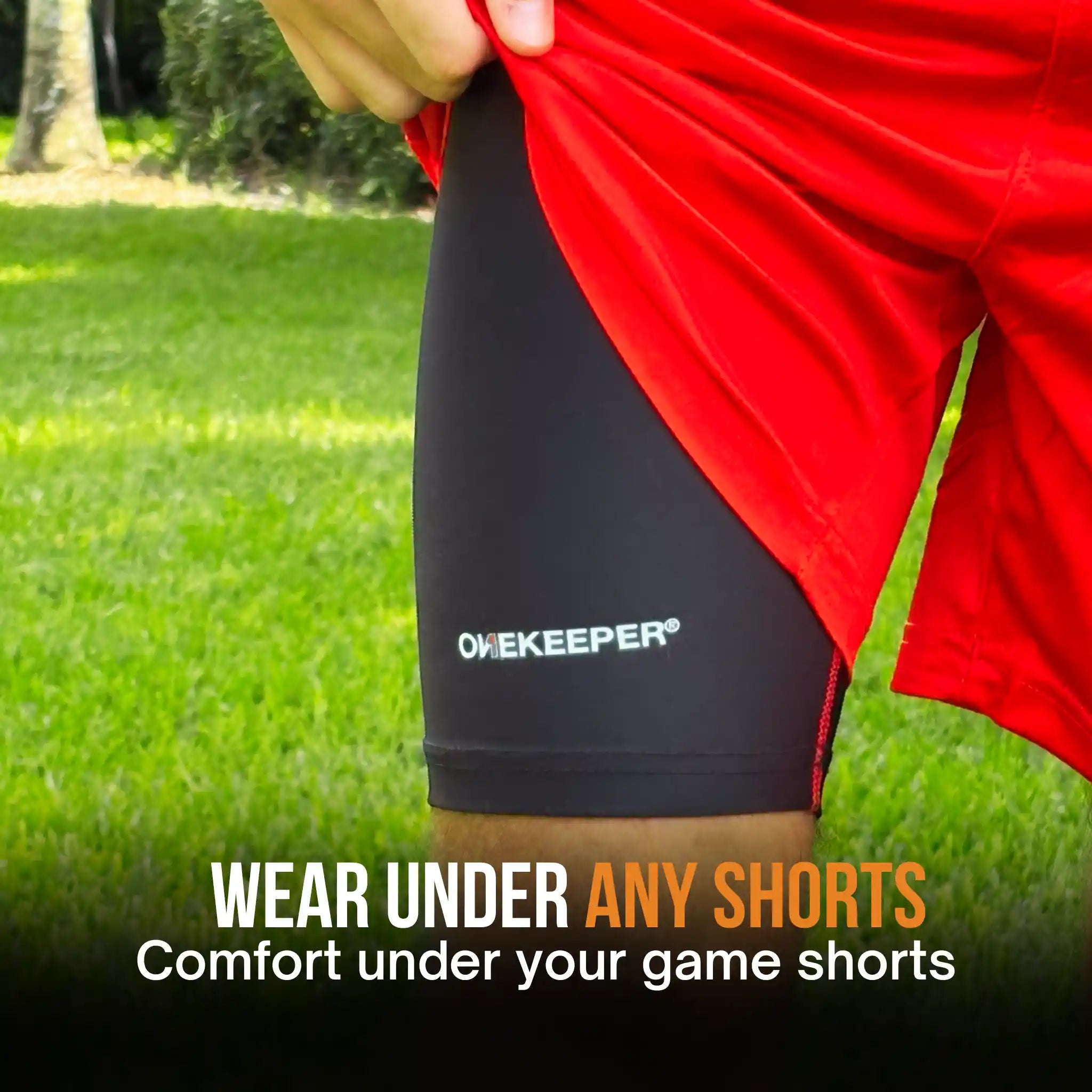 A close-up of a person outdoors on grass wearing black OneKeeper compression shorts under red athletic shorts, with text reading 'WEAR UNDER ANY SHORTS Comfort under your game shorts'.