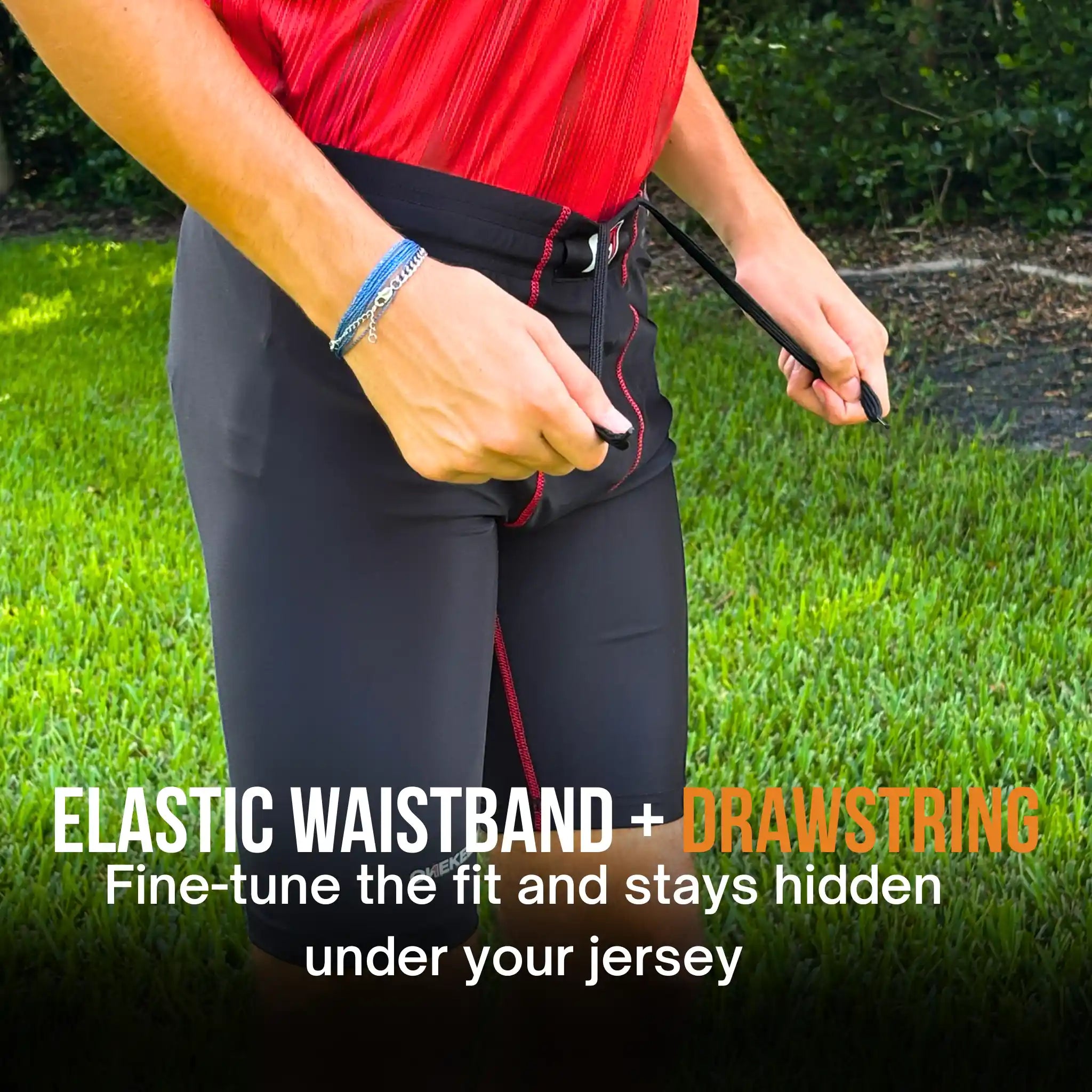 A close-up outdoor shot of a person wearing black onekeeper compression shorts athletic drawstring pants with red stitching, adjusting the waistband with a black drawstring. The person is also wearing a red jersey.