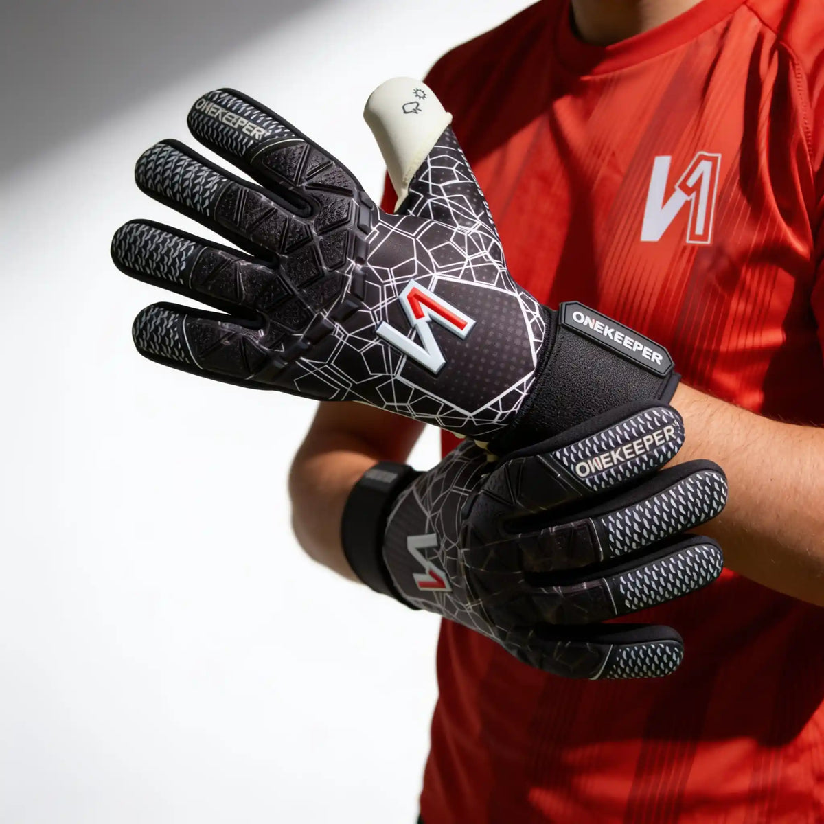 A pair of OneKeeper ctec black soccer goalkeeper gloves being worn by a player in a red jersey. The gloves are black with a white geometric pattern, textured grip panels, and a red and white logo.