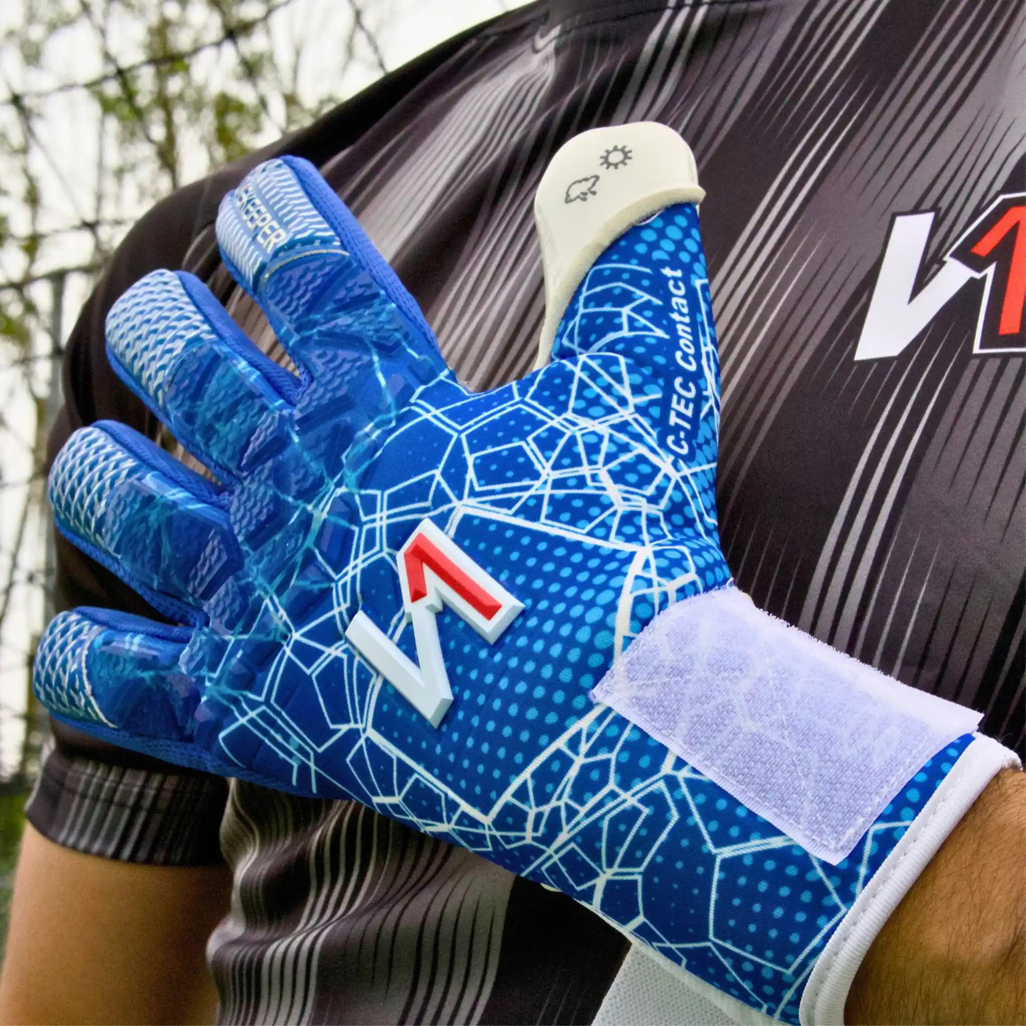 A close-up of a onekeeper ctec blue goalkeeper glove worn on a hand, featuring a blue and white geometric pattern, a white hook-and-loop strap, and a white finger pad, shown against a dark sports jersey.