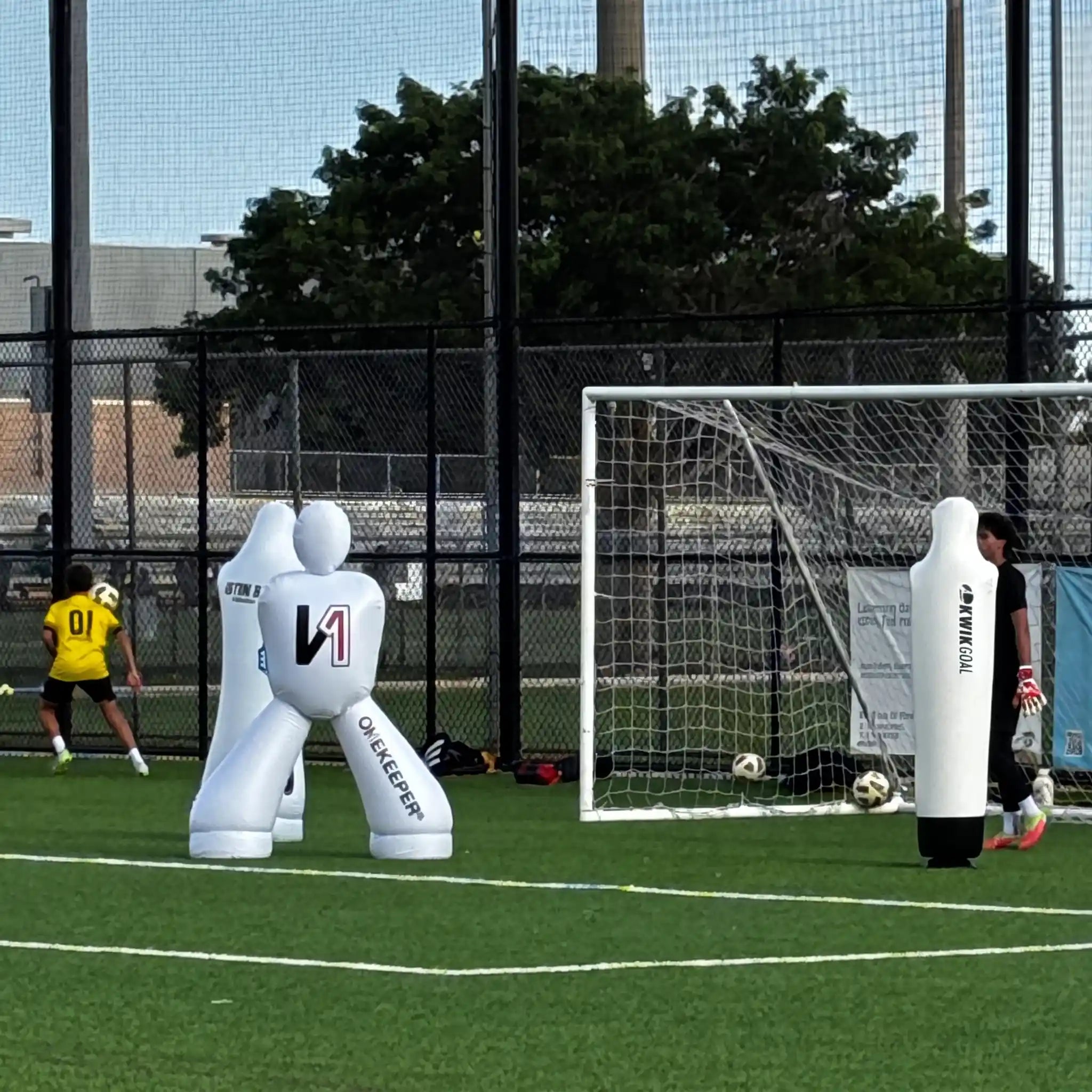 Outdoor soccer field scene with a onekeeper white inflatable goalkeeper dummies, a goal net, a player in a yellow jersey, and trees and fencing in the background.