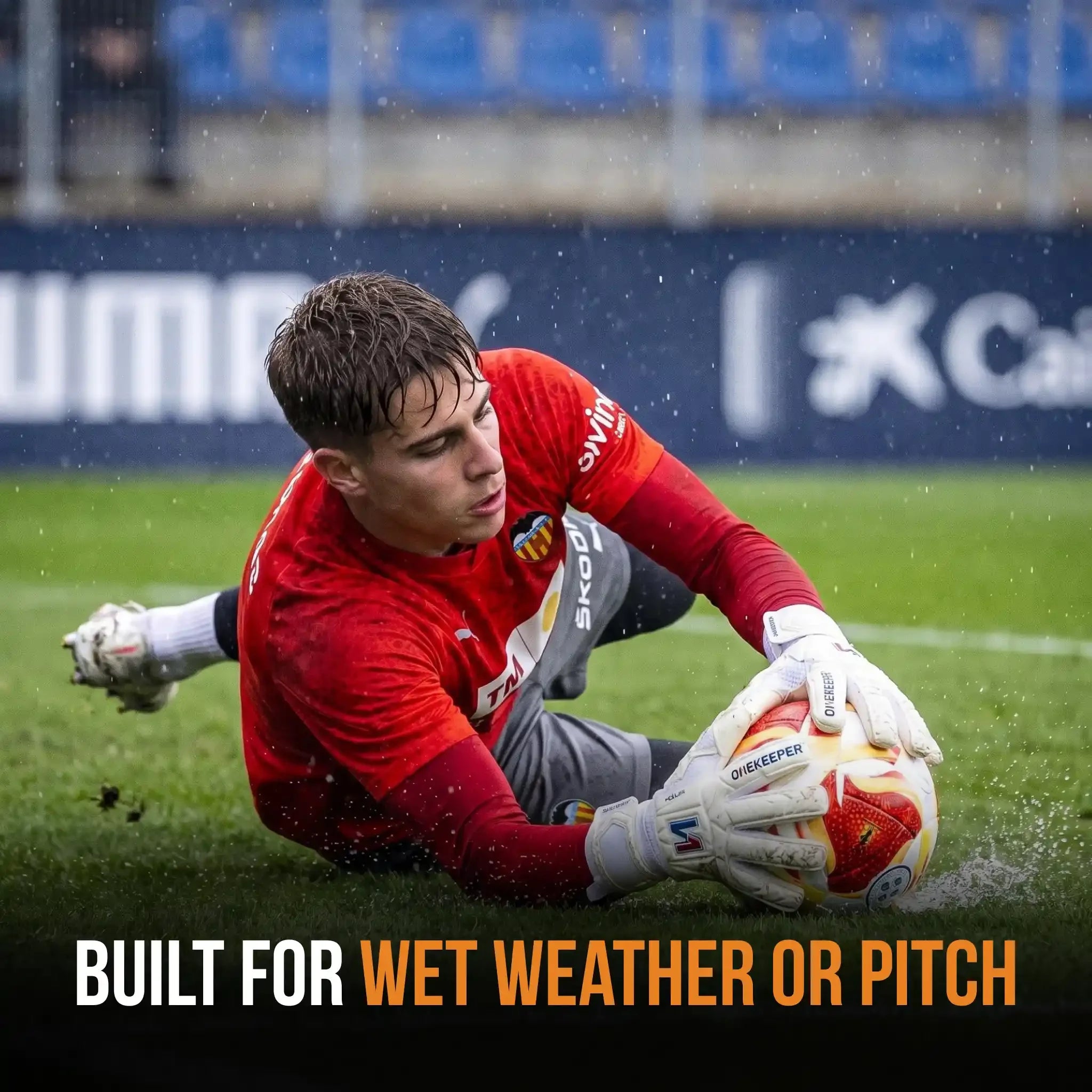 A soccer goalkeeper in a red jersey slides on wet grass while handling a white, red, and orange ball. The image includes the text 'BUILT FOR WET WEATHER OR PITCH' at the bottom.